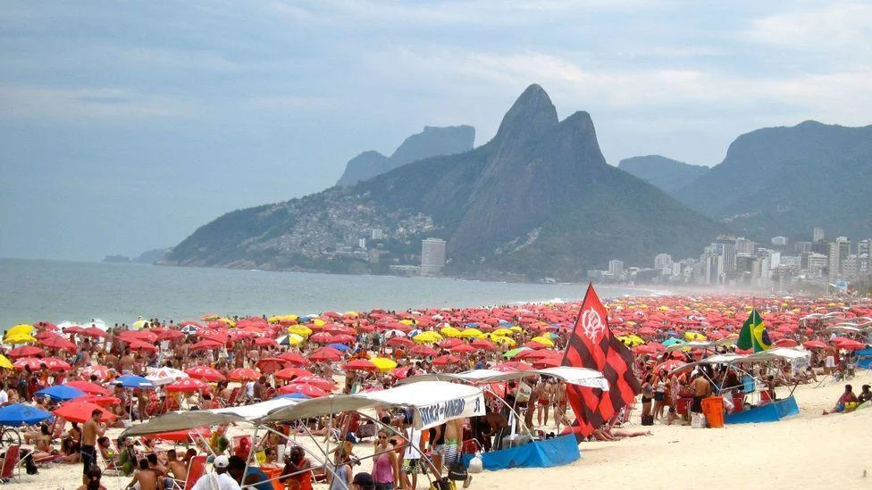Praia de Ipanema lotada com guarda-sóis coloridos e o Morro Dois Irmãos ao fundo